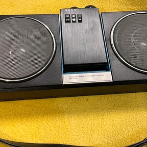 Puede incluir: Un sistema est&eacute;reo port&aacute;til Sony negro con una correa para el hombro. El est&eacute;reo tiene dos altavoces y un reproductor de casetes. El texto en el est&eacute;reo dice "POWERED STEREO STEREO SYSTEM ICF-777W".