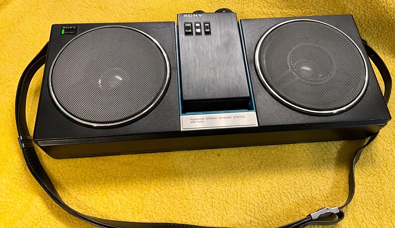 Puede incluir: Un sistema est&eacute;reo port&aacute;til Sony negro con una correa para el hombro. El est&eacute;reo tiene dos altavoces y un reproductor de casetes. El texto en el est&eacute;reo dice "POWERED STEREO STEREO SYSTEM ICF-777W".