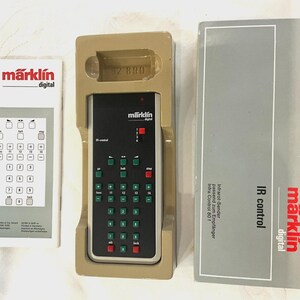 May include: A Märklin Digital IR control remote in a tan plastic tray, accompanied by a white instruction manual and a gray cardboard box. The remote features a black faceplate with green and red buttons, a red slider switch, and white text.