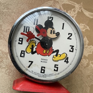 May include: Vintage Mickey Mouse alarm clock with a white face, black numbers, and a chrome bezel. The clock features a red base and a cartoon Mickey Mouse figure. The clock face reads "BAYARD MICKEY" and "WALT DISNEY".