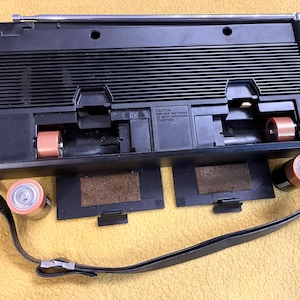 Puede incluir: Reproductor de casetes negro con una antena de radio incorporada, una correa para el hombro y dos compartimentos para bater&iacute;as. El reproductor tiene una cinta de casete negra y marr&oacute;n en su interior. El reproductor est&aacute; sobre una superficie amarilla.