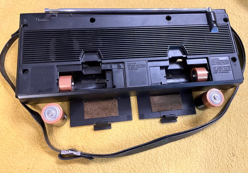 Puede incluir: Reproductor de casetes negro con una antena de radio incorporada, una correa para el hombro y dos compartimentos para bater&iacute;as. El reproductor tiene una cinta de casete negra y marr&oacute;n en su interior. El reproductor est&aacute; sobre una superficie amarilla.