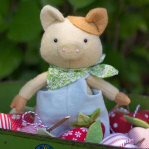 Puede incluir: Un juguete de peluche de cerdo con un mono azul y una bufanda de estampado floral verde y blanco. El cerdo está sentado en un carro verde lleno de bolas de tela con estampado rojo y blanco.