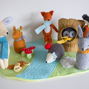 May include: A felt woodland scene with various animal figures, including a deer, fox, hedgehog, and owl. The scene includes a blue river, green grass, and mushroom accents. The animals wear scarves and are arranged on a green felt mat.