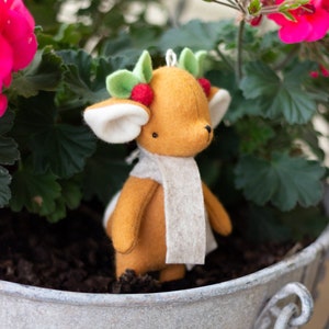 May include: A small, brown felt deer ornament with a white felt scarf and a green holly wreath around its head. The ornament is sitting in a gray metal planter.