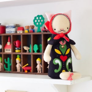 May include: A handmade plush cat doll with a red hooded cape and a black and green patterned dress. The doll is sitting on a shelf with other toys, including a wooden house, a cactus, and a red wagon.