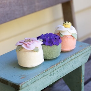 May include: Four felt flower pincushions in different colours and styles. The pincushions are arranged in a row on a light blue wooden surface. The pincushions are made of felt and have a variety of flower designs.
