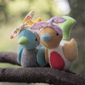 Pode incluir: Dois brinquedos de pássaros de pelúcia com faixas de tecido floral. Um pássaro é azul e cinza com um bico verde, o outro é amarelo e branco com um círculo vermelho e um bico azul.