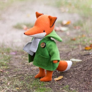 May include: A plush fox toy wearing a green coat with a white scarf and a button. The fox has orange fur and a black nose.