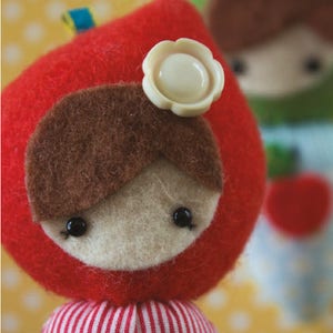 May include: A red felt doll with brown hair, white button eyes, and a white flower with a button in the centre. The doll is wearing a red and white striped dress with a blue bird on the front.