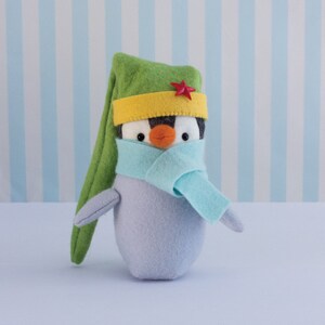 Op de afbeelding: Handgemaakte vilten pinguïnpop met een groene hoed, gele band en rode ster. De pinguïn heeft een lichtblauwe sjaal en een grijze body. De achtergrond is een lichtblauw en wit gestreept patroon.