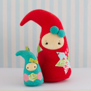 Puede incluir: Dos muñecas de fieltro hechas a mano, una roja con un lazo azul y la otra turquesa con un lazo verde. Ambas muñecas tienen ojos cosidos y detalles de tela de patchwork.