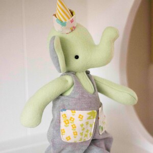 May include: A soft, green elephant plush toy wearing gray overalls with floral pockets and a party hat. The hat is yellow and white striped with a blue pom-pom on top. The toy has gray ears and is standing.