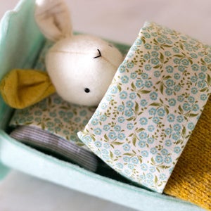 May include: A light blue felt bed with a white bunny plush toy inside. The bed has a floral patterned sheet and a yellow knitted blanket.