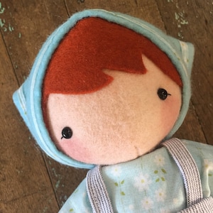 May include: A handmade felt doll with red hair, wearing a blue and white striped hooded shirt and a blue and white floral print dress with black straps.