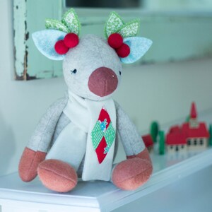 May include: A gray plush reindeer with a white scarf and a red and green patterned scarf. The reindeer has red berries and green leaves on its antlers. The reindeer is sitting on a white shelf.