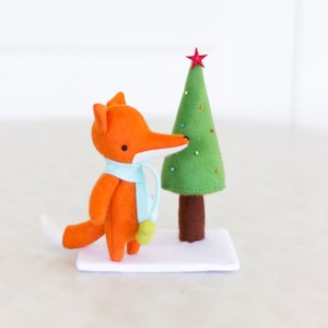 May include: A handmade orange fox plush toy with a white snout and tail, wearing a light blue scarf and holding a green ball. Beside it is a green felt Christmas tree with a red star on top, on a white felt base.