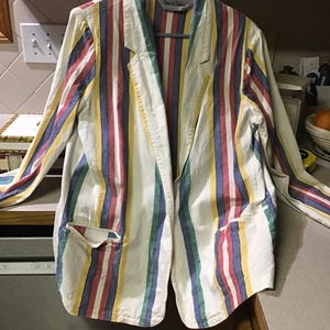 Pode incluir: Blazer vintage com listras verticais vermelhas, azuis, amarelas e verdes sobre um fundo branco. O blazer tem uma gola de lapela clássica, dois bolsos frontais e mangas compridas. O blazer está pendurado em um cabide cinza.