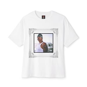 May include: White t-shirt featuring a framed image of a man in a white tank top and bandana. The image is in a decorative frame. The man has a mustache and tattoos. The shirt has a small tag at the neckline.
