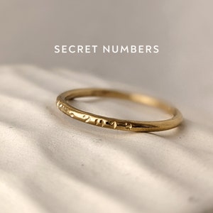 May include: A thin gold band ring with a subtle inscription of numbers around the band. The inscription is difficult to read. The ring is on a white surface. "SECRET NUMBERS" is written in black text above the ring.