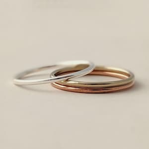 May include: A set of three thin, smooth metal rings stacked together. The rings feature three distinct metallic finishes: one silver-toned, one gold-toned, and one copper-toned. Each band has a simple, rounded profile with a polished, reflective surface.