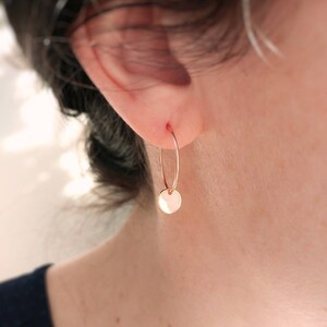 Puede incluir: Un pendiente de aro en oro rosa con un peque&ntilde;o dije circular y plano suspendido del fino aro de alambre. El metal tiene un acabado pulido y reflectante que capta la luz, creando una est&eacute;tica sencilla y limpia para esta pieza de joyer&iacute;a.