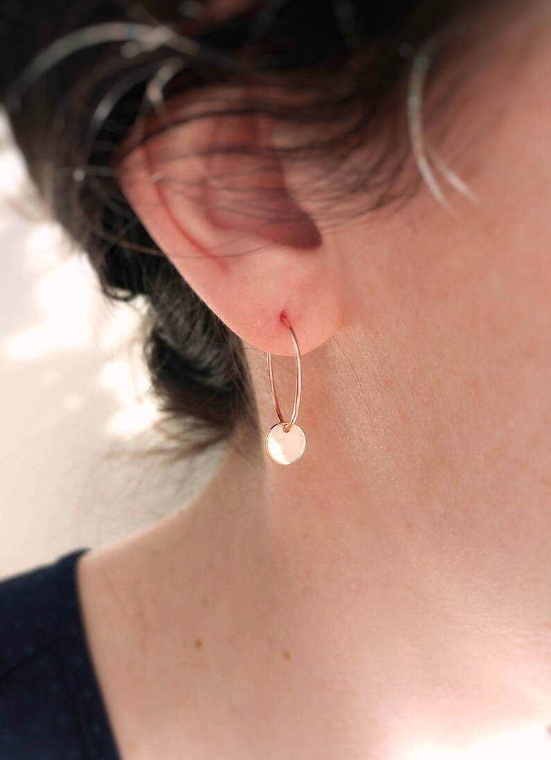 Puede incluir: Un pendiente de aro en oro rosa con un peque&ntilde;o dije circular y plano suspendido del fino aro de alambre. El metal tiene un acabado pulido y reflectante que capta la luz, creando una est&eacute;tica sencilla y limpia para esta pieza de joyer&iacute;a.