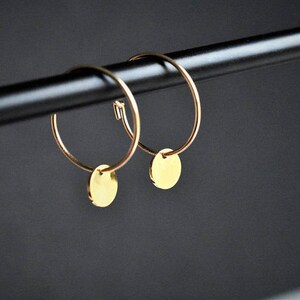 Puede incluir: Un par de pendientes de aro dorados, cada uno con un peque&ntilde;o dije de disco plano y circular suspendido del aro de alambre fino. Los pendientes est&aacute;n fabricados en metal dorado pulido, mostrando un dise&ntilde;o circular sencillo con un acabado de superficie reflectante tanto en los aros como en los colgantes redondos adjuntos.