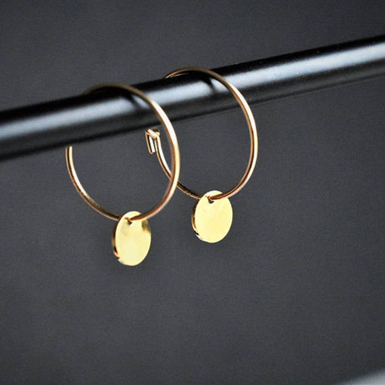 Puede incluir: Un par de pendientes de aro dorados, cada uno con un peque&ntilde;o dije de disco plano y circular suspendido del aro de alambre fino. Los pendientes est&aacute;n fabricados en metal dorado pulido, mostrando un dise&ntilde;o circular sencillo con un acabado de superficie reflectante tanto en los aros como en los colgantes redondos adjuntos.