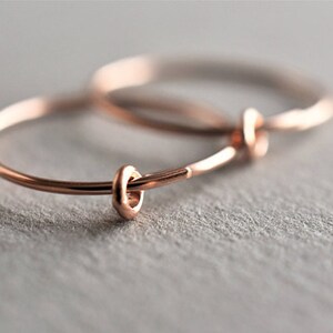Puede incluir: Un par de pendientes de aro circulares y finos hechos de metal en tono oro rosa. Cada pendiente presenta un peque&ntilde;o elemento de anillo liso a juego en tono oro rosa que se desliza a lo largo del fino aro de alambre, creando un dise&ntilde;o sencillo.