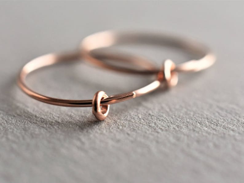 Puede incluir: Un par de pendientes de aro circulares y finos hechos de metal en tono oro rosa. Cada pendiente presenta un peque&ntilde;o elemento de anillo liso a juego en tono oro rosa que se desliza a lo largo del fino aro de alambre, creando un dise&ntilde;o sencillo.