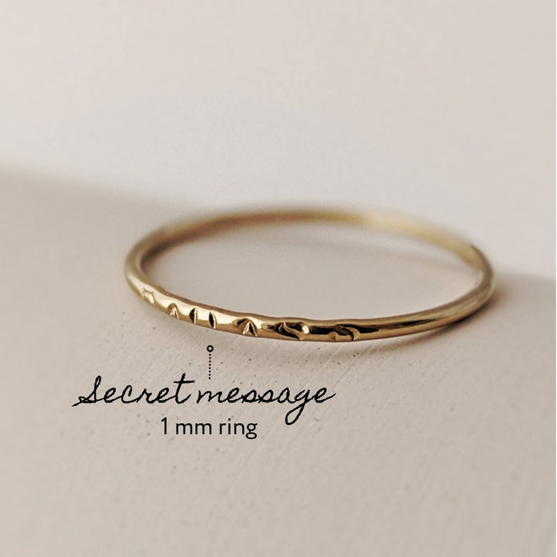 Puede incluir: Un anillo de banda de oro delgado con una superficie texturizada. El anillo tiene 1 mm de ancho y tiene un mensaje secreto grabado en el interior.