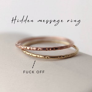 Peut inclure: Deux bagues fines en métal, une en or rose et une en or jaune, présentant une surface texturée avec de petites indentations. Le texte "Hidden message ring" est inscrit au-dessus et "FUCK OFF" est indiqué par une ligne pointillée.