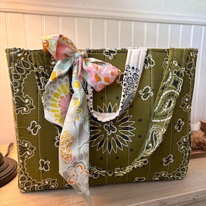 May include: A quilted tote bag featuring an olive green fabric with a white and black paisley bandana print. The bag has two matching fabric handles and is decorated with a tied ribbon in a floral pattern of pink, yellow, and sage green.