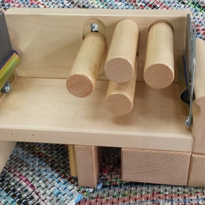 May include: A wooden weaving loom with a light finish. The loom features a series of wooden dowels, a metal reed, and a black handle. The loom is designed for creating woven textiles and crafts.