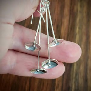 Puede incluir: Pendientes de plata con un diseño único. Cada pendiente presenta tres barras verticales delgadas conectadas a pequeños discos circulares cóncavos. Los pendientes se sujetan con un gancho. Los pendientes están hechos de un metal brillante y reflectante.