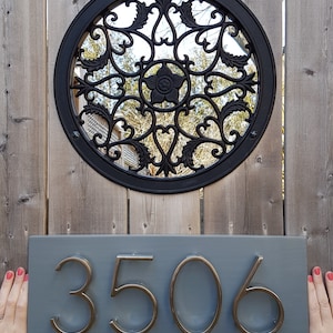 May include: A gray wooden house number sign with the number 3506 in gold metal lettering. The sign is mounted on a wooden fence with a black metal circular design behind it.