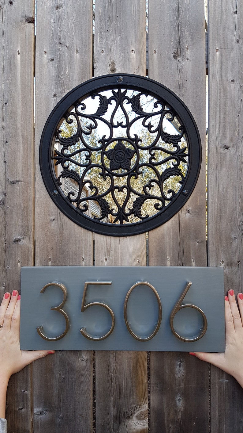 May include: A gray wooden house number sign with the number 3506 in gold metal lettering. The sign is mounted on a wooden fence with a black metal circular design behind it.