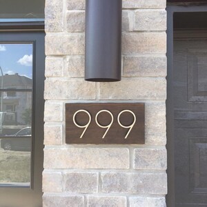 May include: A wooden house number sign with the number 999 in gold metal lettering. The sign is mounted on a brick wall with a black cylindrical light fixture above it.