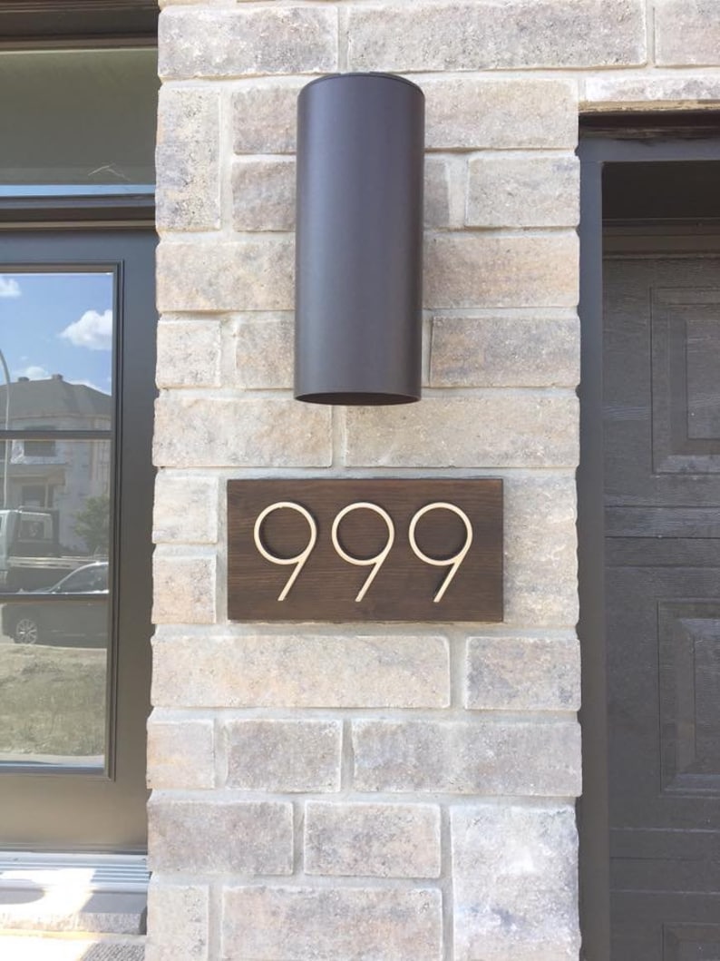 May include: A wooden house number sign with the number 999 in gold metal lettering. The sign is mounted on a brick wall with a black cylindrical light fixture above it.
