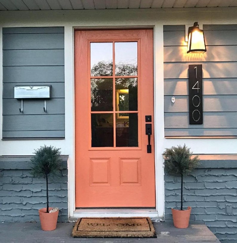 May include: A peach-colored front door with a glass pane and a welcome mat. The door has a black door handle and a black mailbox with a white number "1400" on the side of the house.