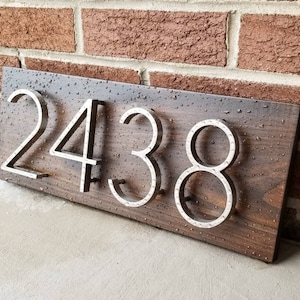 May include: A wooden house number sign with the number 2438 in silver metal letters. The sign is dark brown and has a wood grain pattern. The letters are raised and have a hammered metal finish.
