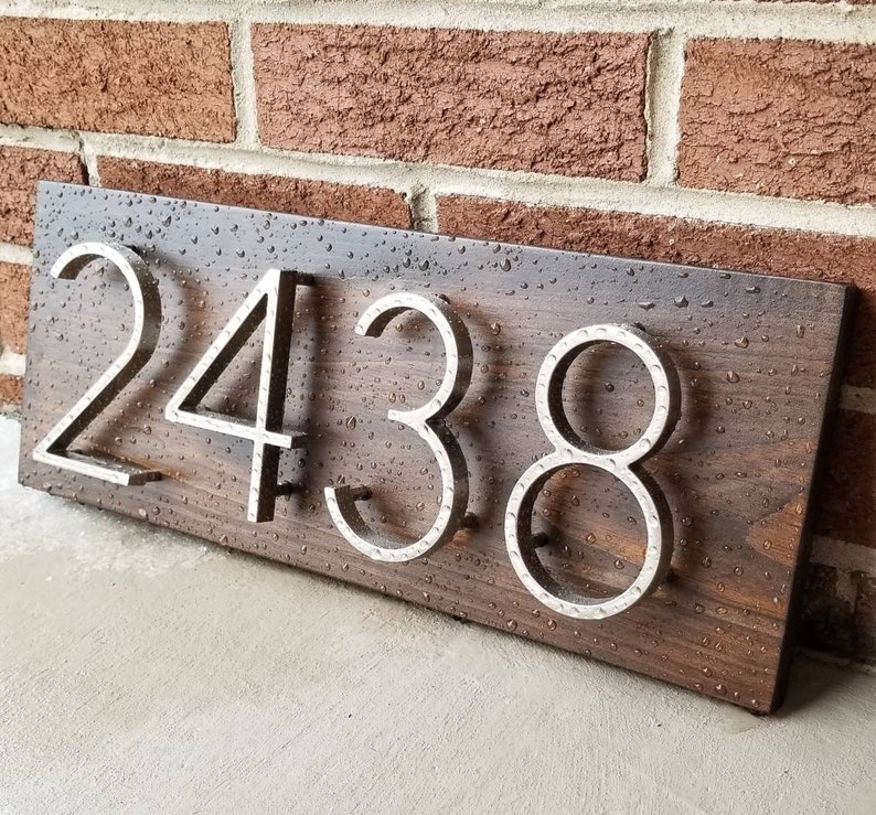 May include: A wooden house number sign with the number 2438 in silver metal letters. The sign is dark brown and has a wood grain pattern. The letters are raised and have a hammered metal finish.