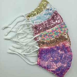 Puede incluir: Cinco mascarillas con elásticos blancos. Las mascarillas están hechas de tela de lentejuelas en varios colores, incluyendo dorado, azul claro, rosa, arcoíris y morado.