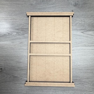 May include: A rectangular light brown wooden frame with a vertical panel design. The frame features a top and bottom ledge, with a horizontal crossbar dividing the interior space into two distinct sections, creating a window-like structure.