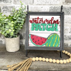 May include: A decorative sign with the words "watermelon patch" in red and green. The sign features a watermelon slice and a whole watermelon. The sign is in a black frame and stands on a wooden surface.