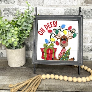 May include: A decorative Christmas sign with a reindeer wearing a scarf and Christmas lights. The sign reads "OH DEER!" and features a gift and Christmas tree. The sign is in a black metal frame.