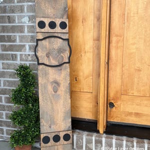 May include: A wooden sign with a black frame and three black circles at the top and bottom. The sign is stained a dark brown color and has a distressed look. The sign is hanging on a brick wall next to a wooden door.