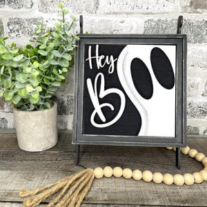 May include: A black and white Halloween sign with the words "Hey Boo" and a ghost design. The sign is in a black metal frame and is displayed on a wooden surface. A small potted plant and a wooden bead garland are also present.