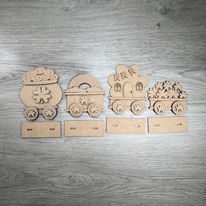 May include: Wooden train set with four cars, each decorated with a different Irish theme. The first car is a pot of gold, the second is a shamrock, the third is a leprechaun's house, and the fourth is a pile of shamrocks.